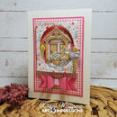 A handmade layered greeting card featuring a pink gingham and floral design, a 3D house cutout with a cat and bird, and a pink ribbon bow at the bottom. The card is displayed on a woven mat with dried flowers against a white wood background.