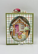 A square decorative art piece or card featuring a painted cottage scene inside an oval frame, bordered by green plaid paper and topped with a pink ribbon loop. The artwork includes a small house with a pink door, flowers, birds, and a cat, with the text 'Simone Carr ART IMPRESSIONS' at the bottom.