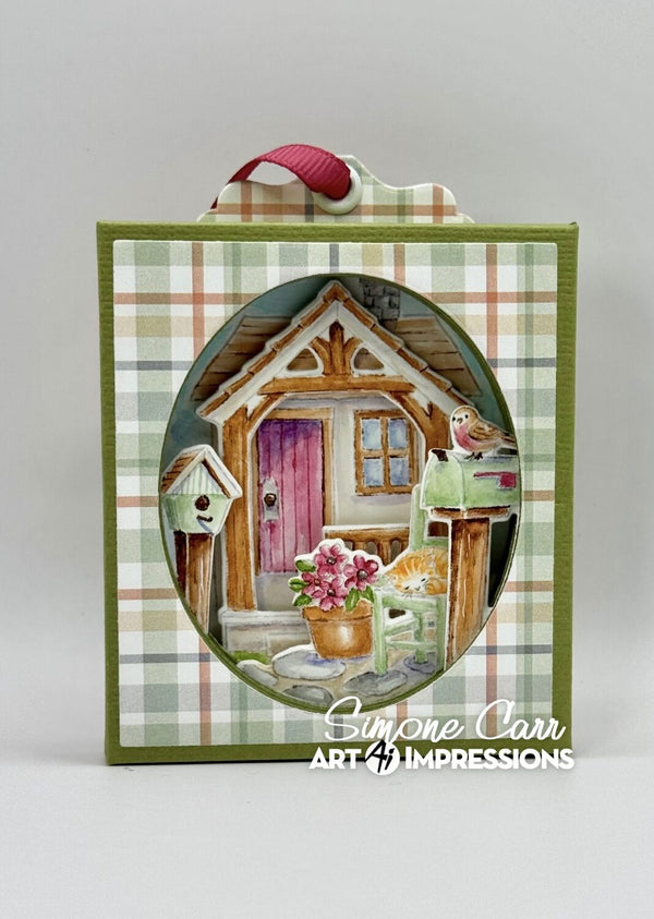 A square decorative art piece or card featuring a painted cottage scene inside an oval frame, bordered by green plaid paper and topped with a pink ribbon loop. The artwork includes a small house with a pink door, flowers, birds, and a cat, with the text 'Simone Carr ART IMPRESSIONS' at the bottom.