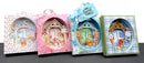 Four decorative gift boxes arranged side by side on a dark surface against a white background. Each box features a colorful illustrated cottage scene visible through an oval cutout, with pastel borders and bows on top.