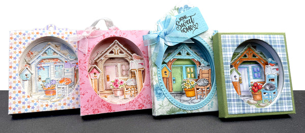 Four decorative gift boxes arranged side by side on a dark surface against a white background. Each box features a colorful illustrated cottage scene visible through an oval cutout, with pastel borders and bows on top.