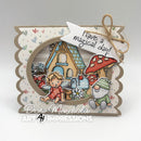 A handmade layered card featuring a scalloped beige border, a circular window view of a whimsical fairy house, a red mushroom, a fairy, and a gnome. A white banner tag reads 'Have a magical day!' tied with twine.