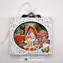 A square decorative art print or ornament with a white floral border and a central oval illustration of a whimsical fairy garden scene. The artwork shows a small fairy, strawberries, a mushroom, a gnome, and a cottage, hanging from a white ribbon.