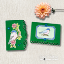 Two small handmade greeting cards with green backgrounds and bird illustrations, displayed on a textured beige fabric background with a peach flower stem. The brand name 'Janet Artman Art Impressions' is visible in the bottom right corner.