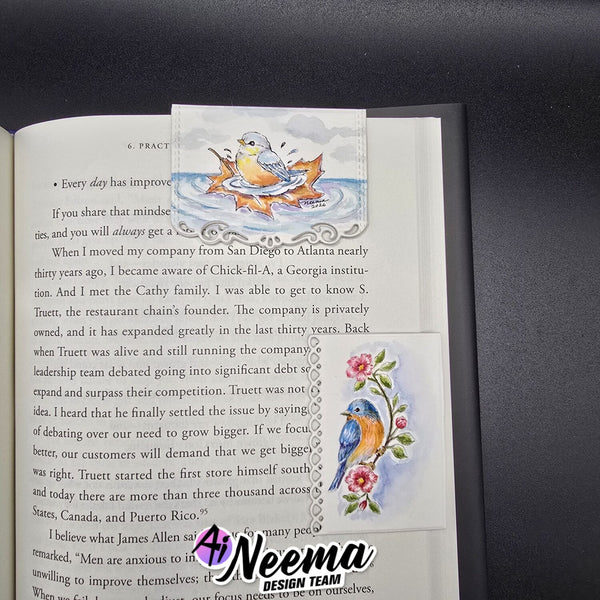 An open book page featuring a decorative printed bookmark or divider with colorful bird and floral illustrations, placed on a dark textured surface. A watermark reading 'Neema Design Team' is visible over the lower right area.