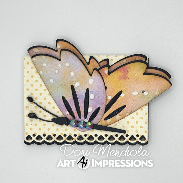A handmade butterfly embellishment with layered pastel wings in lavender, peach, and yellow, mounted on a cream polka-dot card with black scalloped trim. The card is photographed on a light gray background with the text 'Dori Mendiola Art Impressions' printed below.