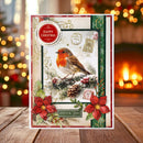 A festive Christmas greeting card featuring a robin bird, red poinsettias, pinecones, and holiday stamps, displayed on a wooden surface with a blurred Christmas tree and fireplace in the background.