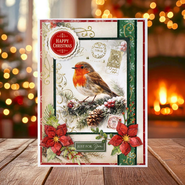 A festive Christmas greeting card featuring a robin bird, red poinsettias, pinecones, and holiday stamps, displayed on a wooden surface with a blurred Christmas tree and fireplace in the background.