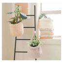 Two woven hanging planters in a natural beige crochet style, one on a black ladder shelf and one hanging from a window sill, each holding a small green plant. A stack of books and a cream basket sit on the windowsill next to a bright window.
