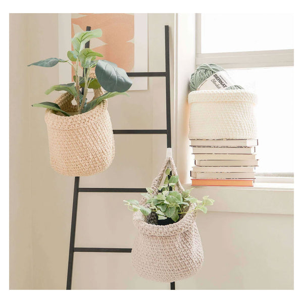 Two woven hanging planters in a natural beige crochet style, one on a black ladder shelf and one hanging from a window sill, each holding a small green plant. A stack of books and a cream basket sit on the windowsill next to a bright window.