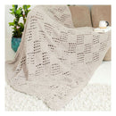 A chunky knit throw blanket in a light taupe-beige color with an openwork geometric pattern, draped over a beige sofa in a living room setting next to a potted plant and a small side table.