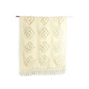 An off-white woven textile with a textured diamond pattern and fringe, hanging from a thin wooden dowel against a plain white background.