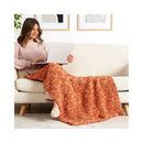 A woman sits on a cream boucle sofa using a laptop, with a chunky rust-orange throw blanket draped across her lap and over the sofa in a bright living room.