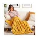 A woman sits on a cream boucle sofa in a bright living room, wrapped in a chunky crocheted mustard yellow blanket. The blanket features a large openwork pattern and is draped over her lap.
