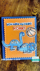 A handmade dinosaur-themed greeting card featuring two blue dinosaurs, a basketball, and the text 'you make my heart ROAR!' on an orange and blue card, photographed flat on a wooden surface.