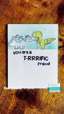 A handmade dinosaur-themed greeting card displayed on a rustic wooden surface. The card features two cartoon dinosaurs, a blue-gray Stegosaurus and a green T-Rex, with the text 'you are a T-RRRIFIC friend'.