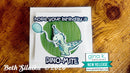 A handmade greeting card featuring a cartoon dino-mite holding a blue balloon, with the text 'hope your birthday is' above and 'DINO-MITE' below, displayed on a wooden surface. A 'gina k.