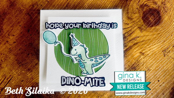 A handmade greeting card featuring a cartoon dino-mite holding a blue balloon, with the text 'hope your birthday is' above and 'DINO-MITE' below, displayed on a wooden surface. A 'gina k.