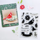 A flat lay of Christmas crafting supplies on a white background, including a handmade card with a red cardinal and the words 'joy to the world!', a sheet of clear stamps with black cardinal, holly, snowflake, and holiday text designs, and several loose decorative paper pieces.