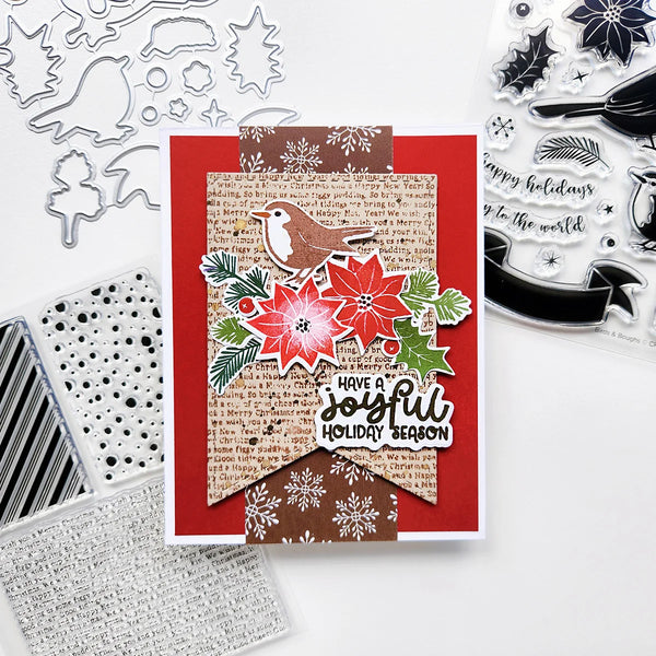 A handmade Christmas greeting card with layered red, brown, and beige paper elements, a bird illustration, poinsettias, holly leaves, and the phrase 'Have a joyful holiday season.' The card is photographed on a white surface with craft dies and clear stamps visible around it.