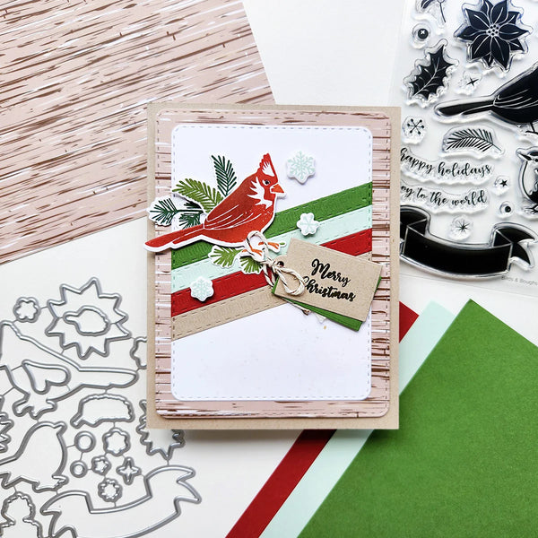 A handmade Christmas greeting card featuring a red cardinal, green and red stripes, snowflakes, pine branches, and a kraft paper tag that reads 'Merry Christmas.' The card is styled on a tabletop with crafting supplies, including clear stamps, metal dies, patterned paper, and colored cardstock.