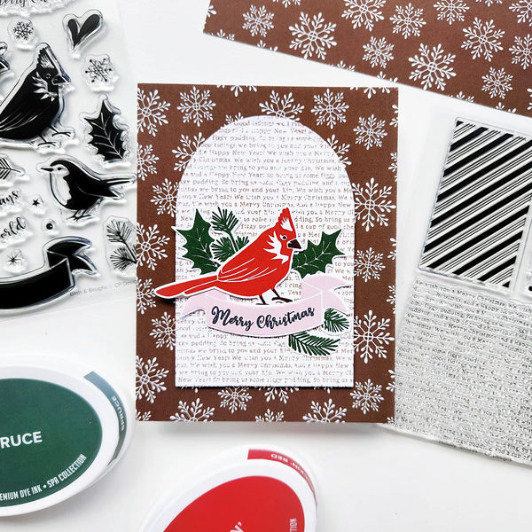 A handmade Christmas card featuring a red cardinal on a brown and white snowflake patterned background, styled with crafting tools and ink pads on a white surface.