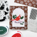 A handmade Christmas card featuring a red cardinal on a brown and white snowflake patterned background, styled with crafting tools and ink pads on a white surface.