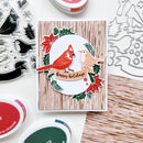 A handmade holiday greeting card featuring a red cardinal and a robin bird on a white banner that reads 'happy holidays', surrounded by red poinsettias, green holly leaves, and a light wood grain background. The card is displayed on a white surface with various crafting tools, including clear stamp sheets, metal die cutters, and ink pads.