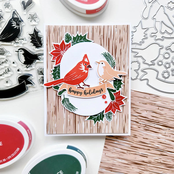 A handmade holiday greeting card featuring a red cardinal and a robin bird on a white banner that reads 'happy holidays', surrounded by red poinsettias, green holly leaves, and a light wood grain background. The card is displayed on a white surface with various crafting tools, including clear stamp sheets, metal die cutters, and ink pads.