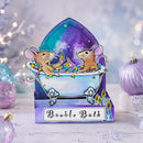 A decorative gift box or card featuring two cartoon mice sitting in a bathtub with bubbles and candles, with the words 'Bauble Bath' on the front, styled on a tabletop with holiday ornaments and soft bokeh lights in the background.