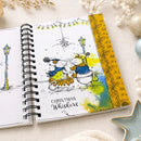 An open spiral-bound notebook or journal lying on a light fabric surface, featuring a whimsical Christmas illustration of two dogs in winter clothing with the text 'CHRISTMAS Whiskers.' The right page has a yellow border with decorative script and leaf motifs.