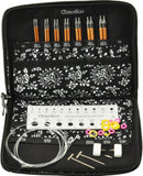 An open black zippered knitting kit case with a black-and-white floral pattern. Inside, six orange-handled knitting needles are stored in the top pocket, and various accessories including a white measuring ruler, metal needles, small rings, and white plugs are arranged in the compartments.