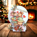 A round, 3D pop-up Christmas card featuring two kissing mice in a sleigh with gifts, set on a wooden table with a blurred fireplace and Christmas tree in the background.
