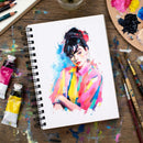 Colorful painting of a woman on a sketchbook with art supplies around