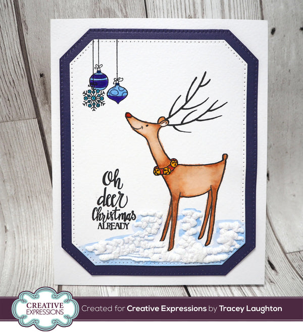 A handmade Christmas greeting card featuring a brown reindeer with antlers, blue hanging ornaments, and the text 'Oh deer Christmas already' on a white card with a navy blue border, photographed on a light wood background.