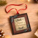 A square Christmas ornament with a red ribbon hanger, featuring a dark brown frame, gold 'Happy Christmas' lettering, and green holly accents on a wooden tabletop background.