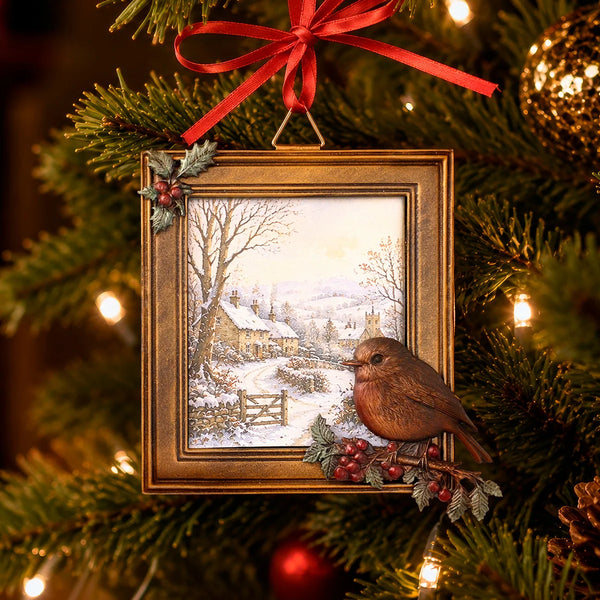 A Christmas tree ornament shaped like a framed winter village scene with a brown bird perched on the lower right, hanging from a red ribbon against a lit Christmas tree background.