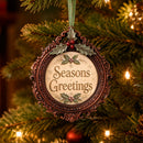 An ornate round Christmas ornament hanging on a lit Christmas tree. The ornament has a dark brown carved frame with holly and red berries at the top, and the words “Seasons Greetings” in brown lettering on a cream center panel.