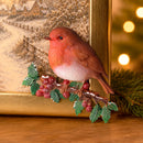 A decorative robin bird figurine perched on a branch with red berries and green leaves, styled in front of a framed winter landscape and holiday lights.