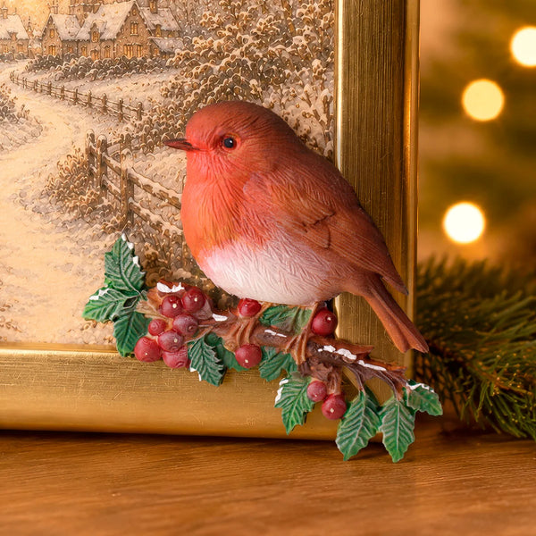 A decorative robin bird figurine perched on a branch with red berries and green leaves, styled in front of a framed winter landscape and holiday lights.