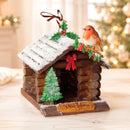 A decorative Christmas log cabin ornament with a small robin perched on the roof, a green tree, holly, red bows, and a plaque reading “New Home,” shown on a tabletop with a softly blurred holiday background.