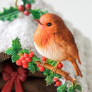 A small decorative robin bird figurine perched on a branch with green holly leaves and red berries, set against a soft white textured background with a red bow visible at the lower left.