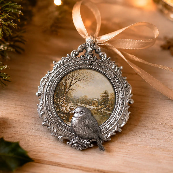 An ornate silver-toned Christmas ornament shaped like a vintage picture frame with a small bird perched on the lower edge, set against a warm holiday background with a ribbon bow and soft lights.