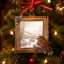 A Christmas tree ornament shaped like a framed winter village scene with a brown bird perched on the lower right, hanging from a red ribbon against a lit Christmas tree background.