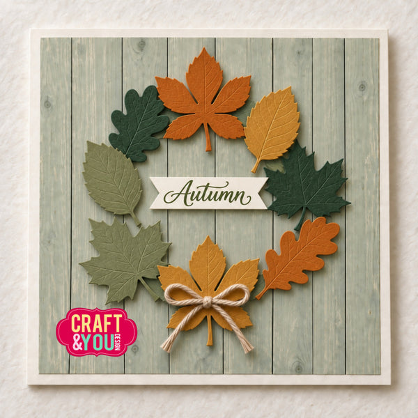 An autumn-themed square card featuring layered paper leaves in orange, yellow, and green arranged in a wreath shape on a faux wood plank background. A white banner in the center reads 'Autumn,' and a pink 'Craft & You' logo appears in the lower left corner.