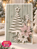 A handmade Christmas greeting card with a rustic light gray wood-plank background, featuring a white laser-cut tree, a pink and white poinsettia, green foliage, pinecones, and a small lantern. The card is displayed in a cozy, festive setting with blurred holiday lights and trees in the background.