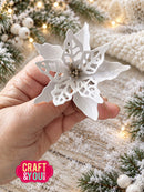 A hand holds a white, snowflake-like poinsettia Christmas ornament against a festive holiday background with pine branches, white berries, and warm string lights. A pink 'CRAFT & YOU' logo is visible in the bottom-left corner.