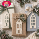Three wrapped gift boxes with pinecones and evergreen branches are arranged on a snowy surface. Each box has a hanging ornament shaped like a house with cutout windows.
