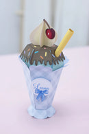 A 3D papercraft model of an ice cream sundae in a light blue and white checkered cone wrapper, featuring brown chocolate sauce, colorful sprinkles, a cream-colored swirl, a red cherry, and a yellow straw.
