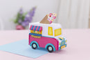 A colorful handmade paper toy ice cream truck parked on a light blue card on a pink surface. The toy features a white cab, a pink side, yellow windows, blue wheels, and a small paper cone on top.